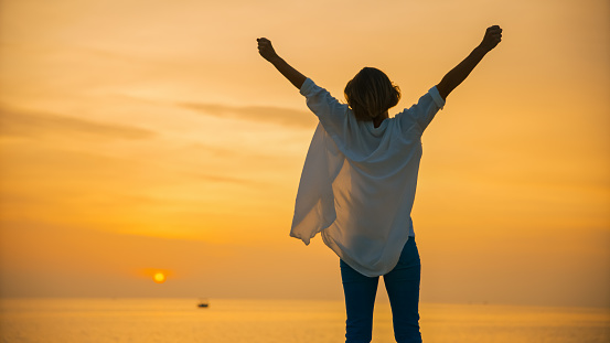 Under the vast orange sky over the Adriatic Sea during sunset in Rovinj,Croatia,a low angle view captures a female tourist standing with arms raised. The scene radiates a sense of joy and wonder,reflecting the magical beauty of the coastal evening