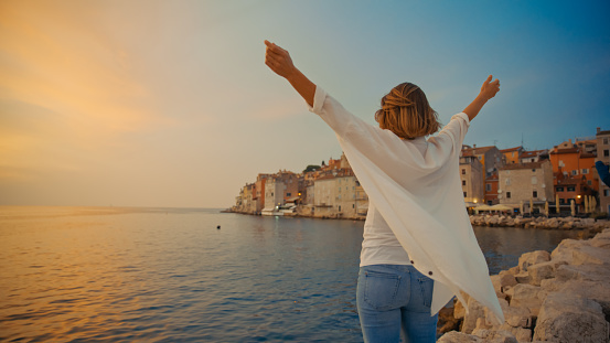 In the enchanting town of Rovinj,Croatia,a rear view captures a carefree woman standing with arms outstretched by the sea against the clear sky and old town during the captivating sunset. The scene reflects the joyous spirit and tranquil beauty of the Adriatic coastal evening