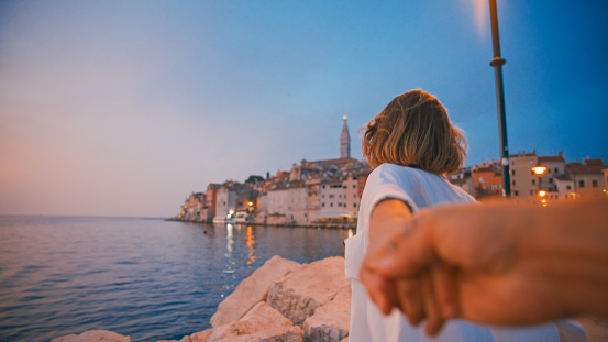 In the romantic allure of Rovinj's dusk in Croatia,a rear view captures a woman holding the cropped hand of a man while standing by the sea against the old town. The scene encapsulates a subtle yet intimate connection,harmonizing with the tranquil beauty of the Adriatic coast