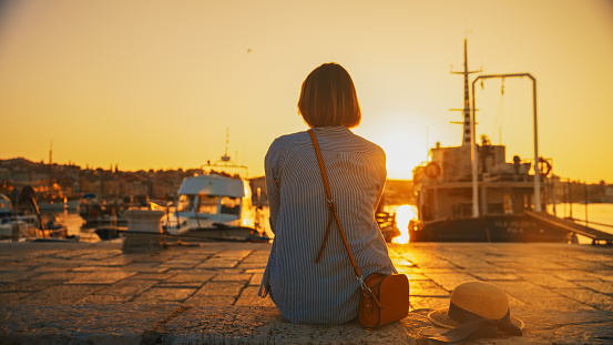 A female tourist,seated on a pier in Rovinj,Croatia,gazes at the idyllic sunset over the harbor,immersing herself in the picturesque beauty of the coastal evening
