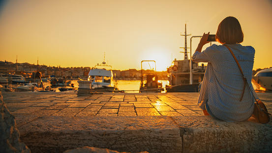 Capturing the moment,A female tourist on a Rovinj pier,engrossed in photographing the enchanting sunset over the harbor using her smartphone,blending modern technology with timeless natural beauty