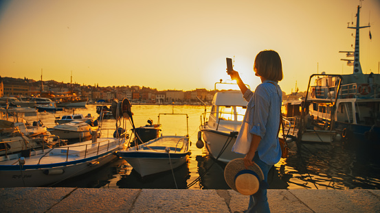 Savoring the sunset,A side view of a female tourist on a Rovinj pier,capturing the picturesque harbor through her smartphone,immersing herself in the beauty of the Croatian sunset