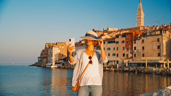 Sunny selfie in Rovinj,A female tourist,adorned with a sun hat,capturing the charm of Rovinj's old town by the sea. The radiant day in Croatia sets the perfect backdrop for a memorable travel moment