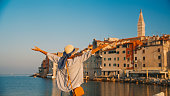 Rear view of female tourist standing with arms outstretched against old town in Rovinj by sea and clear blue sky during summer at Croatia