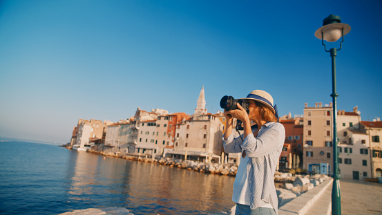 Capturing Rovinj's beauty,A female tourist focused behind her SLR camera,photographing against the clear blue sky and the old town by the sea in Rovinj,Croatia. A blend of technology and artistry in a picturesque coastal setting