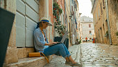 Full length side view of female tourist using laptop while sitting at doorstep of old building by cobbled street at Rovinj,Croatia