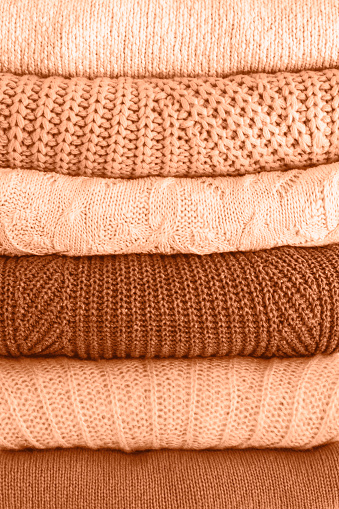 Stack of warm cozy knitted women's sweaters. Cozy autumn or winter clothes. Image toned in trendy color of year 2024 Peach Fuzz. Closeup.