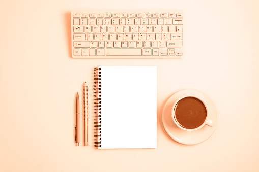 Workplace with computer keyboard, notepad, pen, pencil and cup of coffee or cocoa on peachy background. Top view, flat lay, mockup. Image toned in trendy color of year 2024 Peach Fuzz.