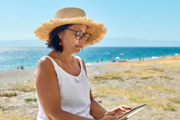 istock Mature woman wearing straw hat using digital tablet at the beach. Middle aged female using digital device for shopping online, banking, social media or reading ebook outdoors. 1845909405