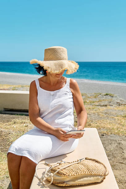 istock Mature woman wearing straw hat using digital tablet at the beach. Middle aged female using digital device for shopping online, banking, social media or reading ebook outdoors. 1845911439
