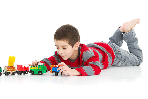 Young boy enjoying playing with a red toy tractor on a dirt path covered with fallen leaves in a forest during a sunny autumn day