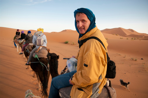 A tourist enjoys a late afternoon camel trek into the Sahara desert. Visible is baggage for overnight stay. A small dog walks alongside. Clear blue African sky