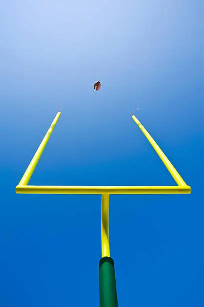 Field Goal or Extra Point in American Football looking up at the goal post as the ball flies through the air against a clear blue sky. Yellow goal post with green padding at the base. 