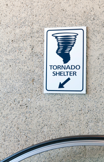 Sign posted on wall in airport above escalator rail indicating with a down arrow where to locate the tornado shelter.  Image of swirling tornado on sign.
