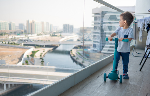 Delightful two and a half year old boy rides his vibrant blue scooter on a terrace with a transparent glass fence, offering a charming perspective of the bustling urban city below