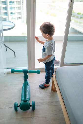 In this heartwarming scene, a cute two and a half year old boy standing by the glass door, clutching his vibrant blue scooter, eagerly expressing his desire to venture onto the terrace for an exciting ride
