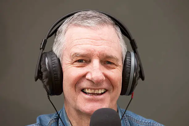 "Smiling while he works, a senior radio DJ and broadcaster talks into his microphone.  He is wearing black headphones and sitting in front of a neutral grey background aa ideal for replacing with a detailed backdrop.  The man has happy eyes and a pleasant friendly face."