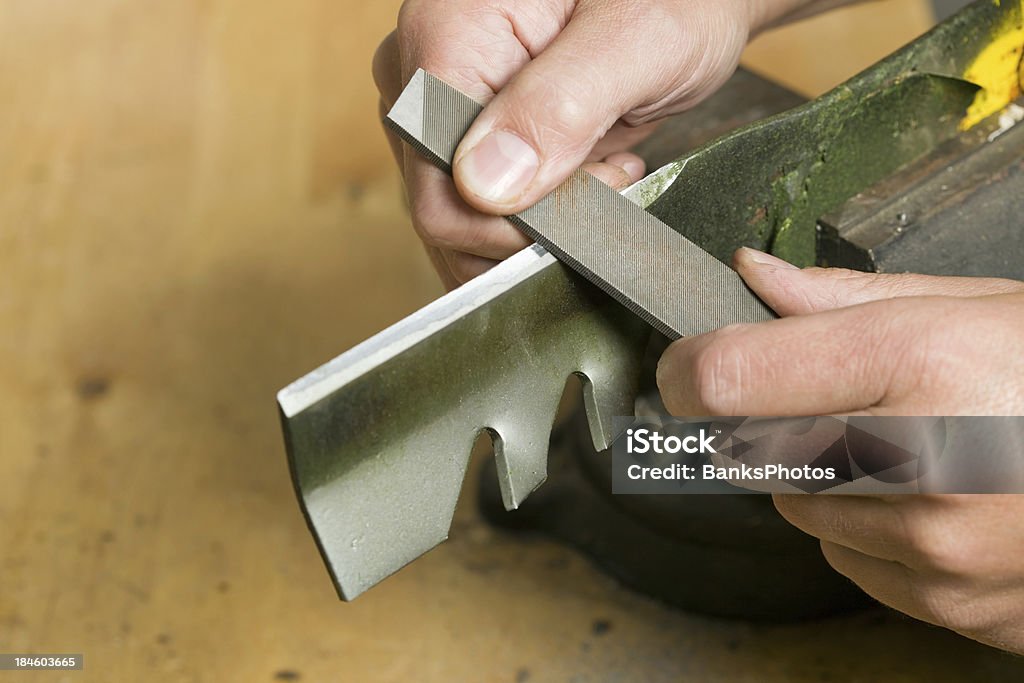 Lawnmower Blade Sharpening with File "Male hands are using a file to sharpen a lawnmower blade which is clamped in a vise atop a wood workbench. Focus is on the file near the bottom hand, the blade is slightly soft." Lawn Mower Stock Photo