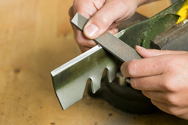 Lawnmower Blade Sharpening with File "Male hands are using a file to sharpen a lawnmower blade which is clamped in a vise atop a wood workbench. Focus is on the file near the bottom hand, the blade is slightly soft." sharpening photos stock pictures, royalty-free photos & images