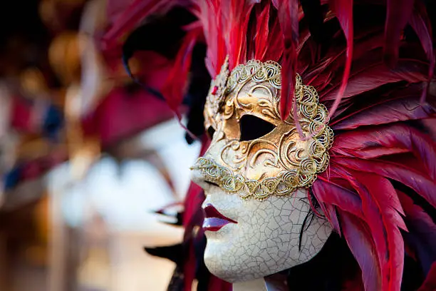 A DSLR photo of a Red Venetian Carnival mask in Venice, Italy. The mask is the main focus of the picture.The mask has pink, purple and red feathers, a golden mask, a porcelain face with texture and cracks and red lips. Other blurred masks are in the background of the picture.