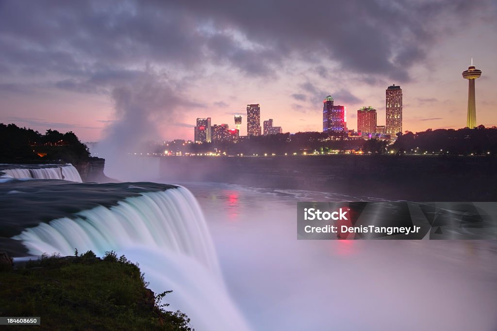 Niagara Falls Flowing Niagara Falls from the American side with the skyline of the city of Niagara Falls in the background at dusk. The Niagara Falls is one of the largest and most famous waterfall in the world.  Niagara Falls consists of three waterfalls that straddle the international border between Canada and the United States. New York State Stock Photo