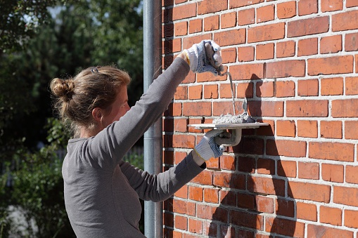 Female house owner working on the brick wall. Replacing old mortar between bricks with new to keep the house healthy.