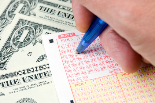 A male hand is using a pen to mark lottery numbers on a ticket resting on a background of one dollar bills.A related image from my portfolio:aA