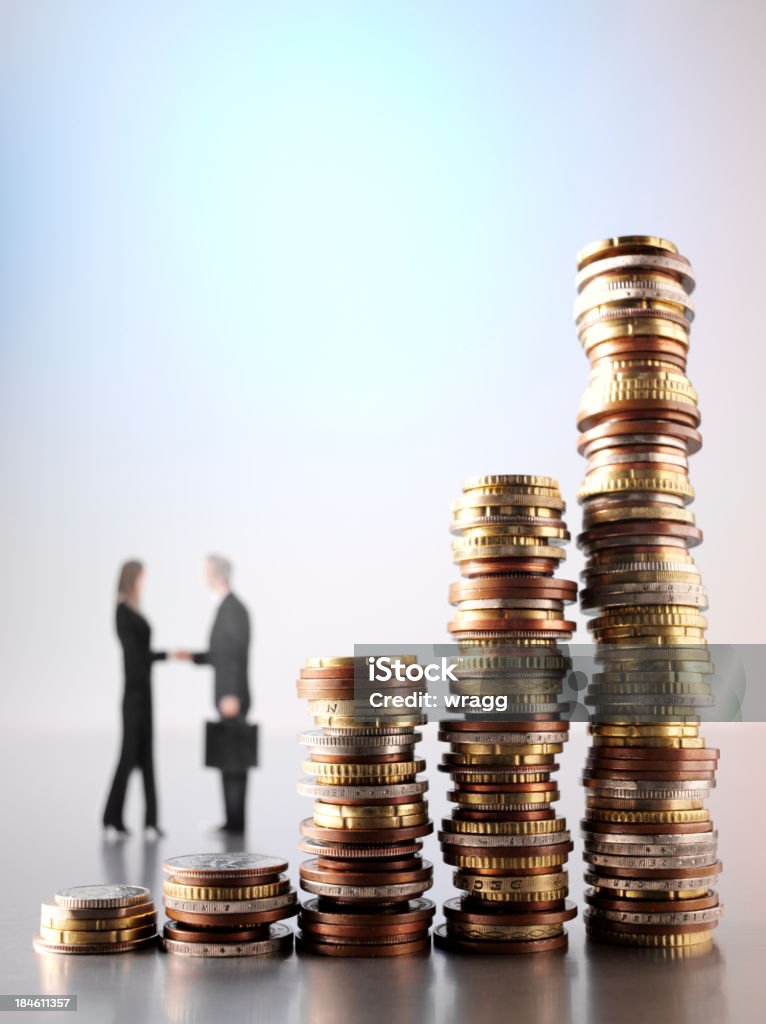 Economic Agreement Economic agreement over a towers of coins on stainless steel with business people shaking hands in the background. Differential focus and copy space Currency Stock Photo