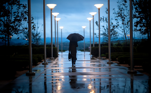 Unidentifiable businessperson walking on sidewalk with a black umbrella on a rainy day at dusk