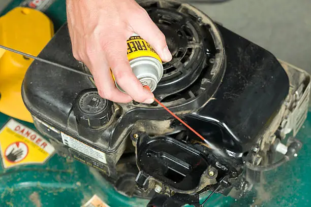 "A male hand is using spray cleaner on the carburetor linkage of a gas lawnmower engine. Focus is on the hand, the spray tip and engine are slightly soft."