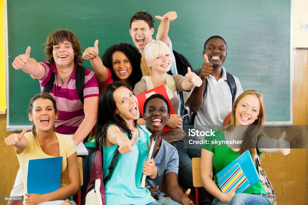 Cheerful students looking at the camera with their thumbs up. Front view of teenage students sitting in front of the blackboard with their thumbs up and looking at the camera.    High School Student Stock Photo