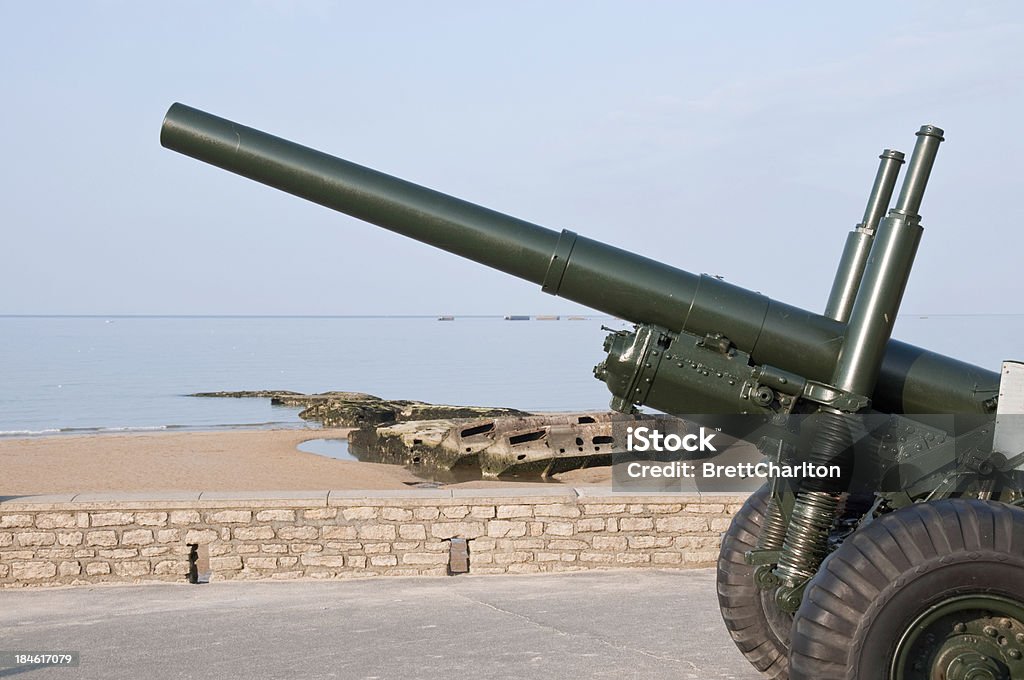 D-Day Gold Beach Military field gun stands at the head of Arromanches  beach - Code name Gold Beach. In the background the remains of the fameous floating 'Mulberry' harbour Bridge - Built Structure Stock Photo
