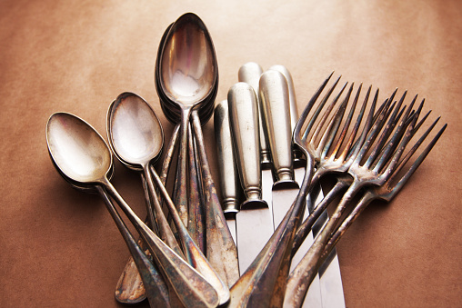 A studio shot of vintage silverware on brown background paper background. Spoons, knifes and forks grouped by type arranged in fan shape. From above.