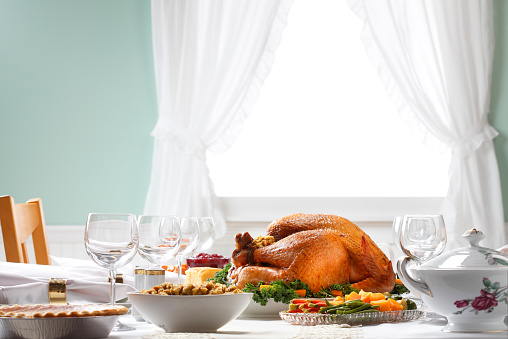 A table setting prepared for a traditional Thanksgiving dinner.  A beautiful, bright natural light fills the room from the picturesque window in the background.  With the turkey as the centerpiece of the table, this spread reflects bounty and the quintessential Thanksgiving experience.  