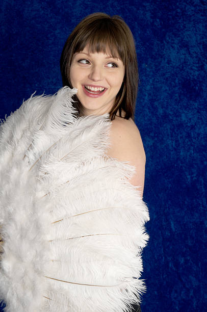 Laughing Burlesque dancer with fan. Young woman hiding behind large white fan laughs at someone off camera.  Vertical studio shot on blue crushed velvet. ostrich feather stock pictures, royalty-free photos & images