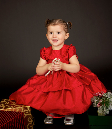 A cute two year old girl with pigtails and a red dress. She is holding candy canes and has Christmas Presents around her.