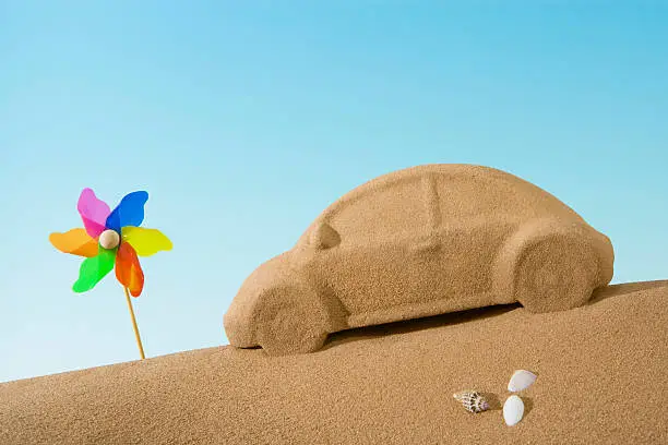 A sand sculptured car is sitting on a sandy beach with a few small shells, which are white and brown.  In front of the car is a rainbow-colored windmill.  The background is a light blue gradient.  There is a light shining on the sand, highlighting the detail of the sculpture.