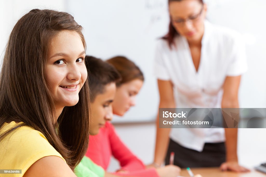 Smiling  Teenage Girl In The Classroom Teenage girl looking at camera and smiling in a classroom with her classmates and teacher in the background. Adult Stock Photo