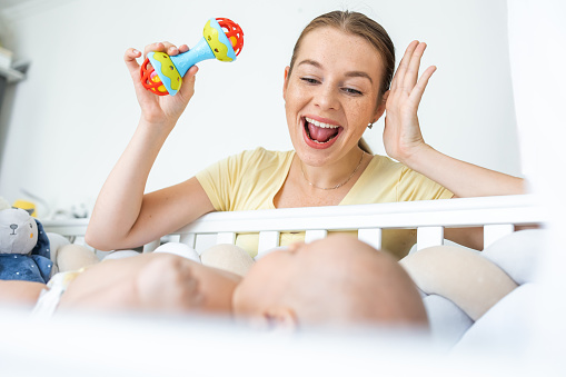 Young mother shaking plastic colorful rattle toy while playing with her baby at home