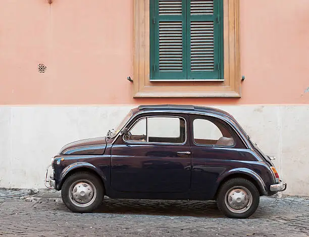 Side view of a small, classic Italian car (a vintage Fiat 500) on a cobbled backstreet in Rome.