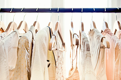 A row of light-colored clothing secured on wooden hangers is visible from about armpit level up, dangling from a rod which stretches from one end of the frame to the other.  Behind the rack, two large windows brighten the room with daylight.