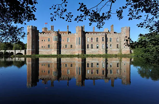 "A beautiful and historic 15th century English castle reflected in it's moat and framed by trees and pond plants in semi silhouetteHerstmonceux, East Sussex, UK"