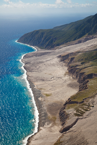 The remains of recent pyroclastic flows of the Soufriere Hills Volcano form the east coast of Montserrat. Aerial view from helicopter. You can see fresh ash being carried away by the waves. The cliff visible to the right is the old coastline while the island is growing steadily.