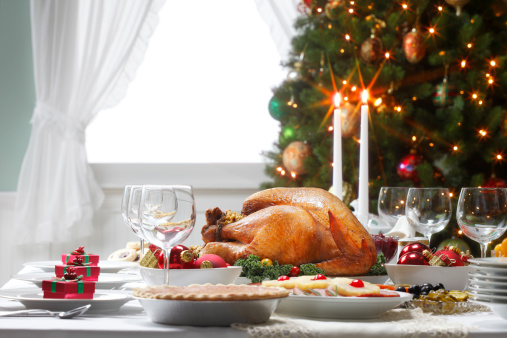 Close up of a turkey at a traditional Christmas dinner.  The natural light pours in from the inviting window framed by a decorated Christmas tree in the background.  Lit with candles, the feast is ready for its guests to indulge in a quintessential Christmas meal filled with turkey and pie.