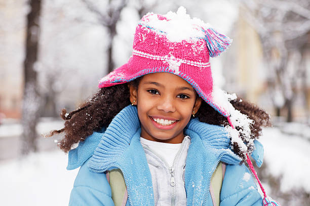 Beautiful African-American girl looking at the camera. Happy girl surrounded by snow with snow covering her hat. kids winter coat stock pictures, royalty-free photos & images