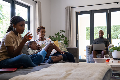 An African family enjoy a lazy afternoon sitting on the sofa watching television together while their father is sitting alone working on his laptop computer.
