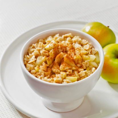 Healthy breakfast of oatmeal and apples; white bowl on white plate. Small yellow apples soft focus in background.