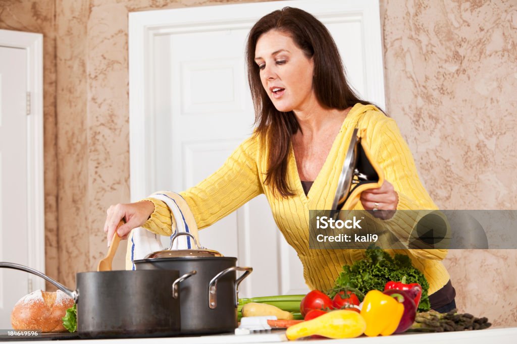 Woman in kitchen cooking on stove "Mature woman (40s) in kitchen by stove with fresh vegetables, cooking dinner." Cooking Pan Stock Photo