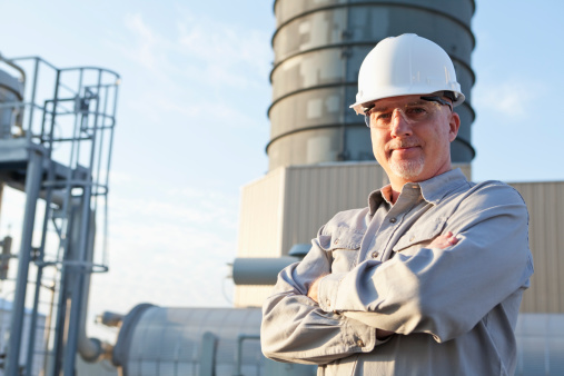 A serious, mature man, in his 50s, working at a power station, standing outdoors with his arms folded, wearing a gray uniform, white hardhat and safety glasses.  We see the manufacturing facility in the background.