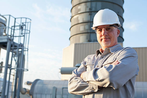 Engineer wearing hardhat at industrial facility A serious, mature man, in his 50s, working at a power station, standing outdoors with his arms folded, wearing a gray uniform, white hardhat and safety glasses.  We see the manufacturing facility in the background. hard hat stock pictures, royalty-free photos & images